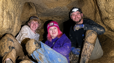 Three people in a cave, wearing helmets and muddy clothes, smiling.