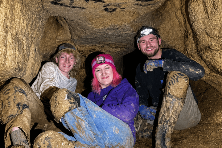 Three people in a cave, wearing helmets and muddy clothes, smiling.