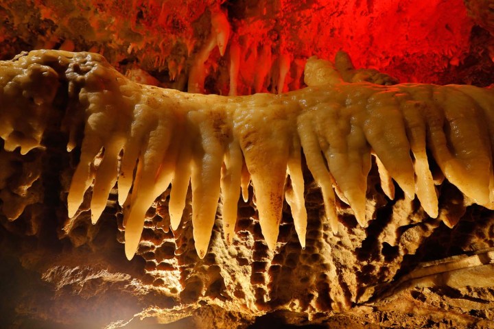 Stalactites hanging from a cave ceiling, illuminated by red and white lights.